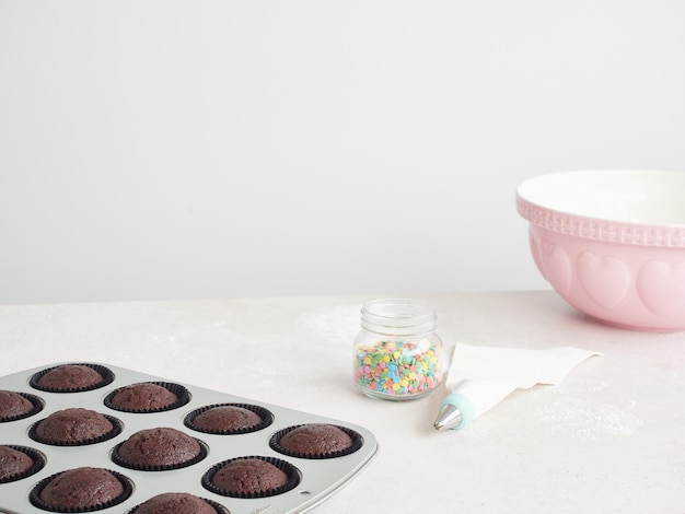 집에서 굽고 컵케이크 장식 베이킹 트레이에 초콜릿 컵케이크 조리대 위에 노즐과 분홍색 그릇이 있는 식용 색종이 파이핑 백