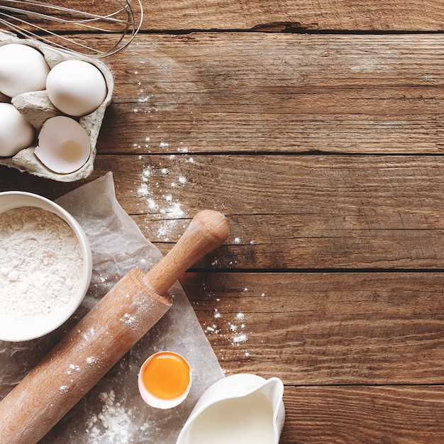 Baking ingredients, kitchen utensils on old wooden background. Cooking dough, preparing egg yolk, flour, rolling pin, milk, parchment paper, whisk, salt, sodium. Concept flat lay photo with copy space
