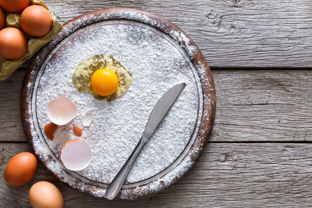 Bakken concept. Bestrooid meel en eieren op houten snijplank in de buurt van mes, ingrediënten koken. Bereid je voor op het maken van gistdeeg. Bovenaanzicht