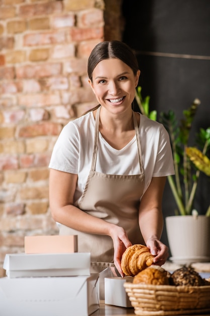 Bakken, verpakken. Vrolijke glimlachende nette vrouw in beige schort croissant aanbrengend vak verpakking producten in patisserie