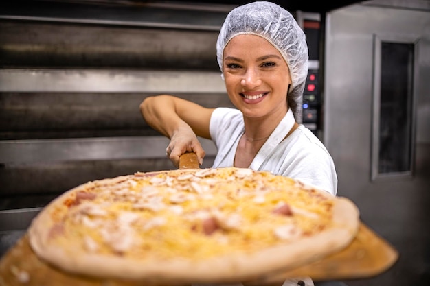 bakker die bij de oven staat en dienblad vasthoudt met versgebakken pizza in de bakkerij voor voedselproductie