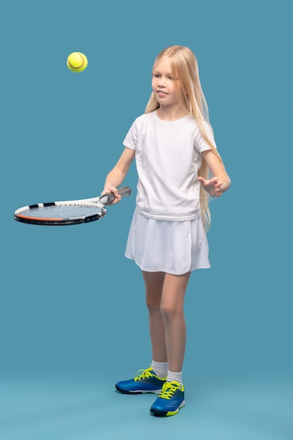 Bal gooien. Glimlachend betrokken meisje in witte sportkleding die bal gooit met tennisracket die handen uitoefent
