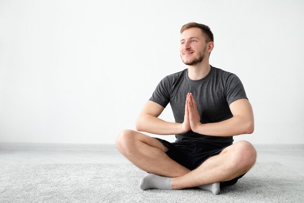 Equilibrio yoga uomo felice meditazione domestica mente pacifica ragazzo sorridente in pantaloncini tshirt seduto posa loto namaste mani sul pavimento luce parete interno sfondo spazio copia