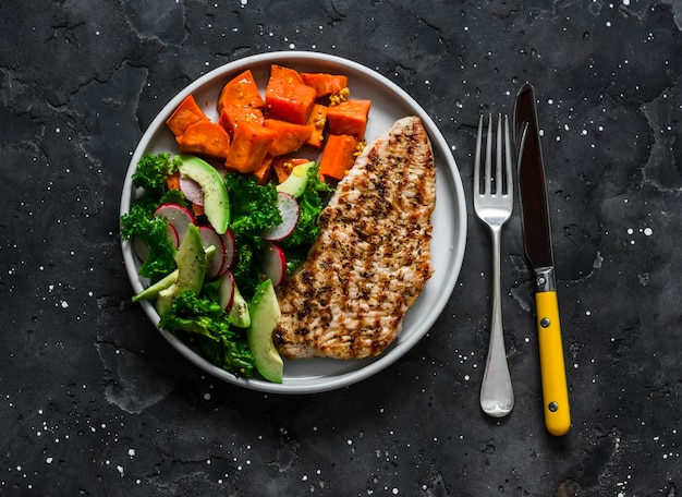 Balanced diet lunch grilled turkey boiled sweet potatoes kale cabbage avocado cucumbers radish salad on a dark background top view