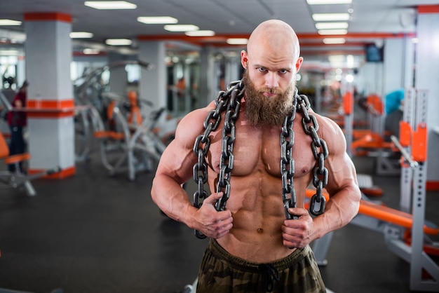 A bald bearded bodybuilder with large relief muscles holds a heavy thick chain in his hands