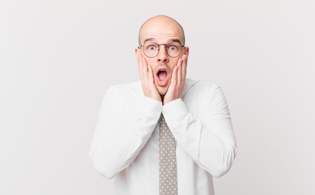 Bald businessman feeling shocked and scared, looking terrified with open mouth and hands on cheeks