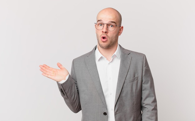 bald businessman looking surprised and shocked, with jaw dropped holding an object with an open hand on the side