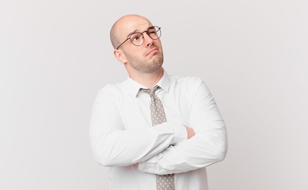 Bald businessman shrugging, feeling confused and uncertain, doubting with arms crossed and puzzled look