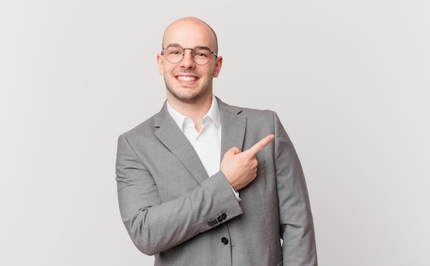 Bald businessman smiling cheerfully, feeling happy and pointing to the side and upwards, showing object in copy space