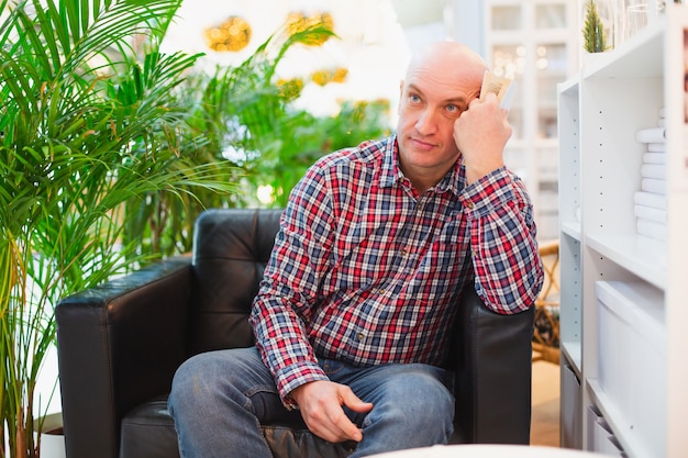 Bald european man in a red checked shirt and blue jeans sits in an armchair in a bright apartment with green plants in the background, thoughtful with a book in his hands