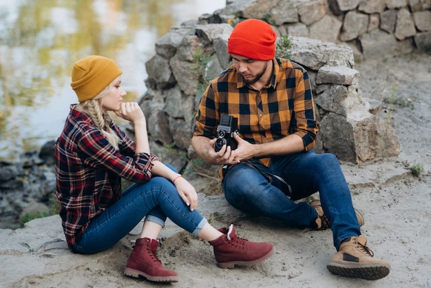 Bald guy with a beard and a blonde girl in bright hats on the background of the river are taking pictures with an old camera