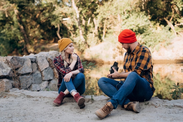 Bald guy with a beard and a blonde girl in bright hats on the background of the river are taking pictures with an old camera