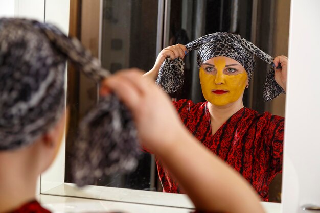 A bald jewish woman on her face with a cosmetic mask ties a\
scarf on her head in front of a mirror