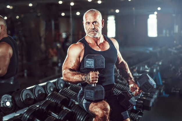 Bald male bodybuilder holds dumbbells in arms while posing in gym in dark concept of bodybuilding