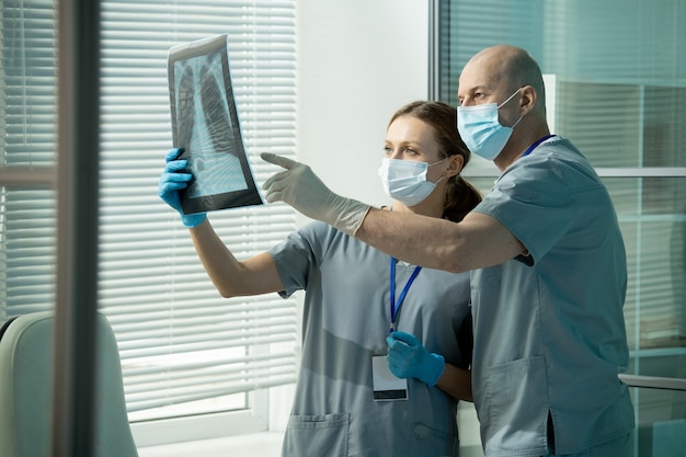 Bald male clinician in uniform and protective mask and gloves pointing at lung x-ray held by his assistant during discussion of pathologies