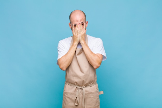 Bald man covering face with hands, peeking between fingers with surprised expression and looking to the side