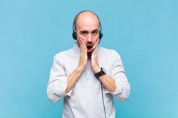 Bald man feeling shocked and scared, looking terrified with open mouth and hands on cheeks