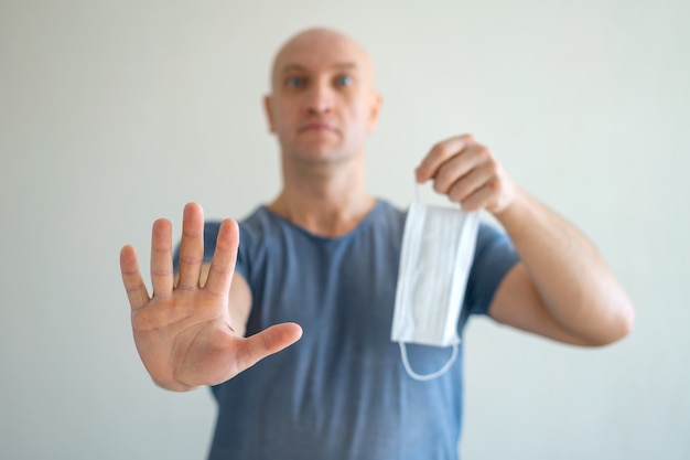 A bald man holds a medical mask in his hands, his other hand held out in protest