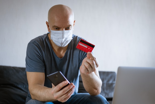 Bald man in a medical mask with a credit card sits on the couch at a laptop with a phone