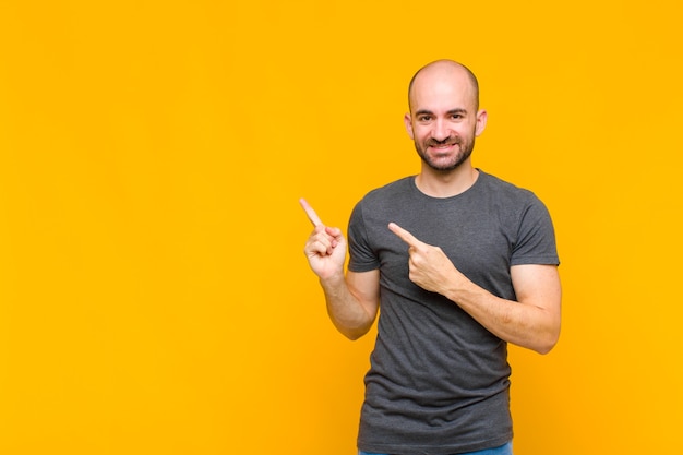 Bald man smiling happily and pointing to side and upwards with both hands showing object in copy space