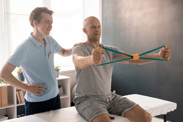 Bald man stretching resistance band during exercise for arms while sitting on medical couch, his physiotherapist standing near by and helping him