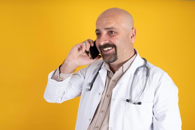 A bald man in a white medical doctor coat and wearing stethoscope talking on a phone