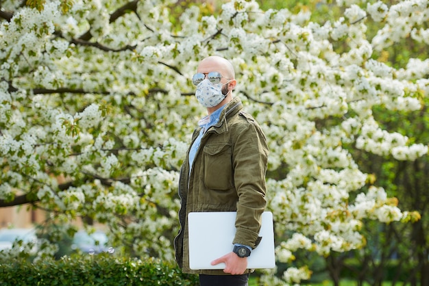 コロナウイルスの蔓延を防ぐために医療用フェイスマスクにひげを生やしたハゲ男が、公園のラップトップを持って歩きます。ある男は、花の咲く木々の間の庭でn95フェイスマスクとパイロットサングラスをかけています。