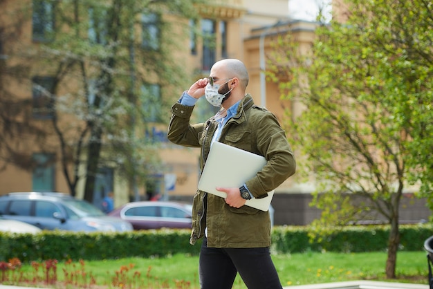 A bald man with a beard in a medical face mask to avoid the spread coronavirus walks with a laptop in the park. A guy wears n95 face mask and a pilot sunglasses on the street of the city.