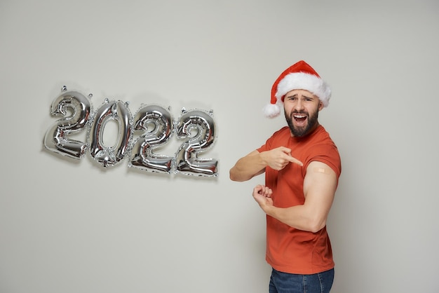 A bald man with a beard in a Santa hat is screaming and showing biceps with a plaster after vaccination against Coronavirus (Covid-19) near foiled silver balloons in the shape of 2022.