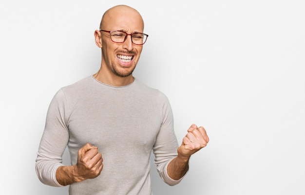 Bald man with beard wearing casual clothes and glasses excited for success with arms raised and eyes closed celebrating victory smiling. winner concept.