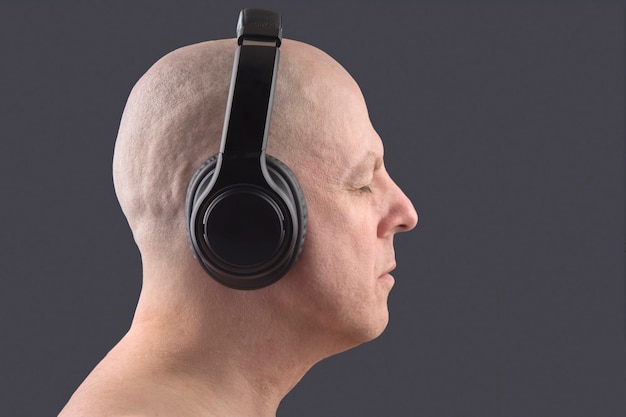 Bald man with closed eyes listens to music with headphones on dark background