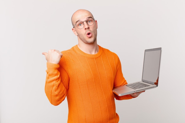 Bald man with computer looking astonished in disbelief, pointing at object on the side and saying wow, unbelievable