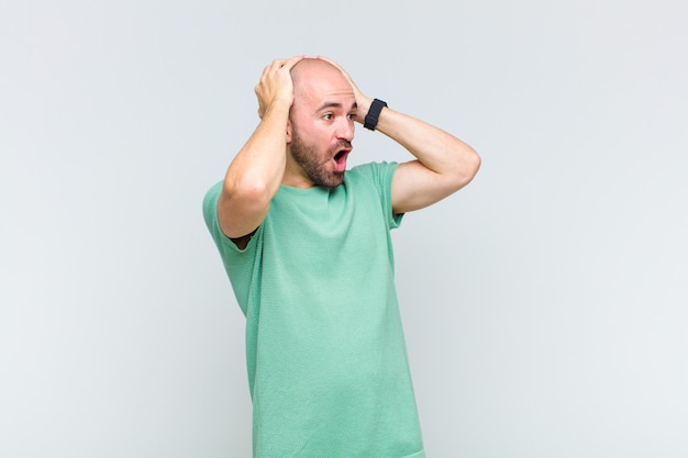 Bald man with open mouth, looking horrified and shocked because of a terrible mistake, raising hands to head