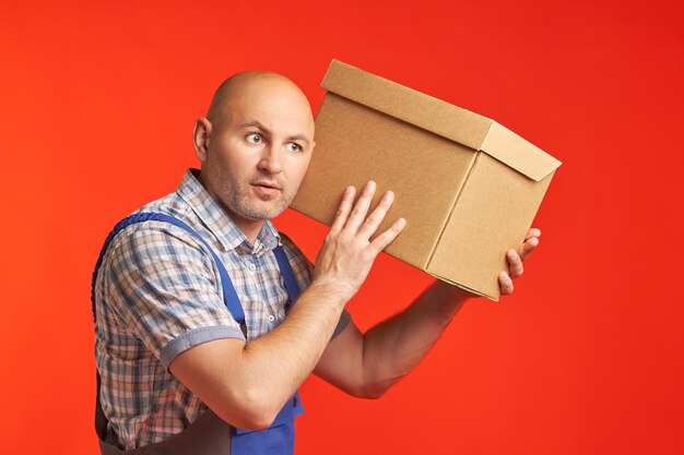 Bald man in work clothes holds a box near his head with surprise