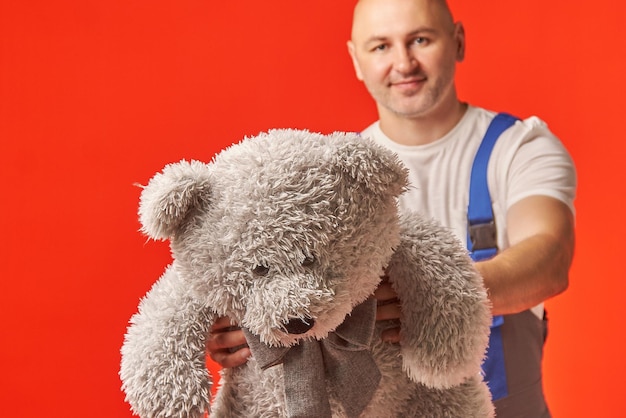 Bald man in work clothes holds a gray teddy bear on a red background