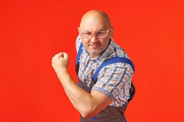 Bald man in a work suit and goggles shows muscles on a red background