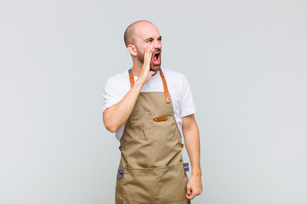 Bald man yelling loudly and angrily to copy space on the side, with hand next to mouth