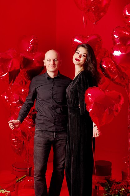 Bald man and young woman in black stand with heart-shaped balloons