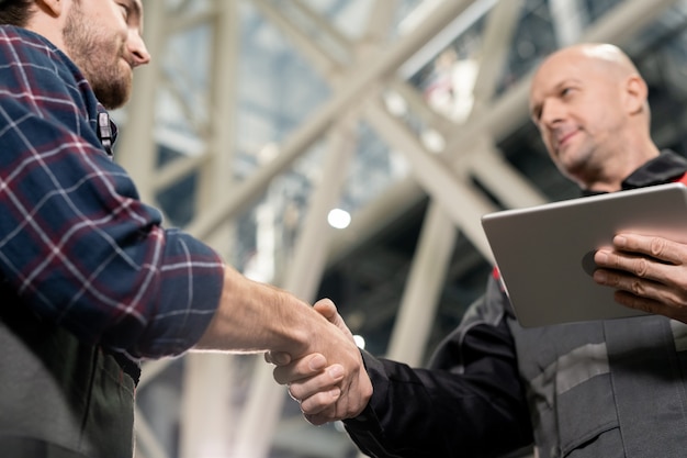 Photo bald mature foreman with digital tablet greeting his young subordinate by handshake before meeting in warehouse or factory environment