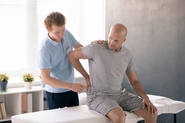 Лысый зрелый мужчина трогает болезненную спину, сидя на медицинской кушетке в реабилитационных клиниках и слушая рекомендации врача