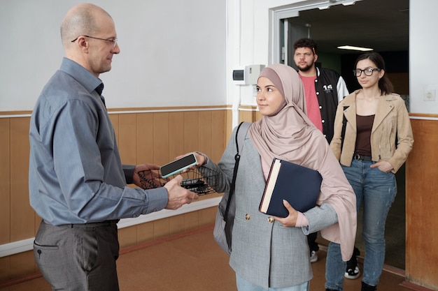 Professore maturo calvo dell'università in abbigliamento casual intelligente che tiene il cestino mentre gli studenti mettono i loro smartphone lì prima della lezione