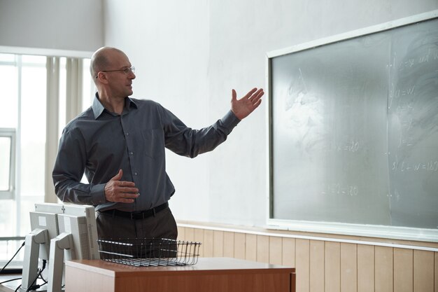 Лысый зрелый учитель смотрит и указывает на заметки на доске во время объяснения