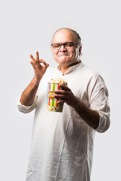 Bald senior Indian asian man eating popcorn over white wall with cheerful expressions