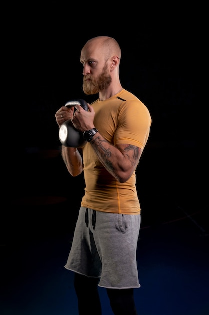 Bald tattooed athlete with red beard holds a kettlebell with two hands