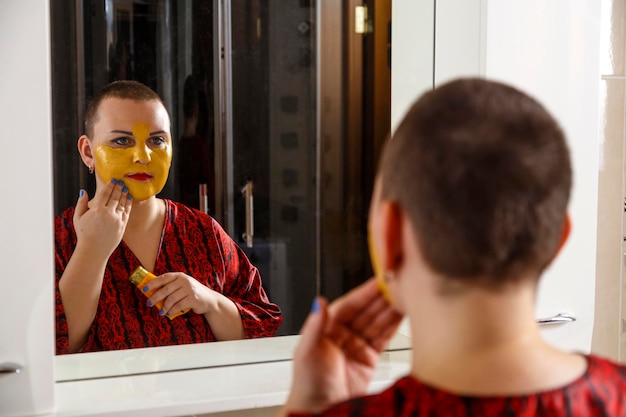 A bald woman applies a cosmetic mask on her face in the bathroom Reflection in the mirror