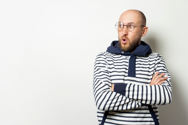 Bald Young man with a beard in glasses of a jacket with a hood looks away with a surprised face on an isolated white. Gesture of shock, surprise