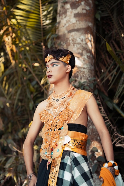 A Balinese man with a golden costume and makeup on his face is standing in front of the coconut tree