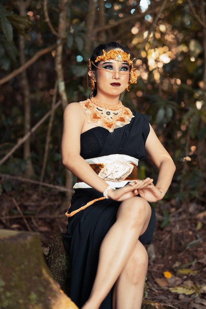 Balinese woman sitting beautifully on the rock in a black dance costume with makeup on her face and a gold crown on her head