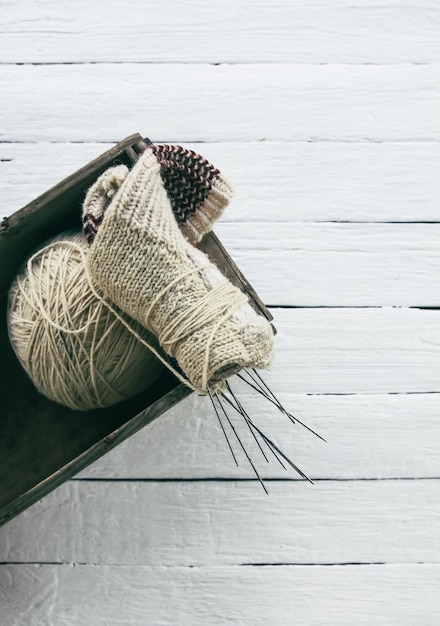 Ball of rough woolen yarn and knitting needles in a box on white wooden boards background.