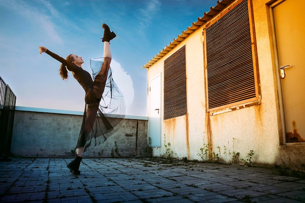 Ballerina in a black skirt and bodysuit does vertical splits on the roof of a building against the sky